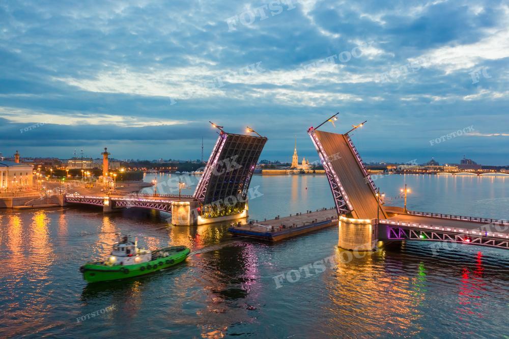 На фотографии изображён Дворцовый мост в Санкт-Петербурге во время разводки. Вечернее небо и освещение добавляют сцене уникальную атмосферу, а отражение огней в водах Невы создаёт волшебный эффект. Такое фото может использоваться в туристических брошюрах, культурных публикациях, репортажах о городе и как элемент урбанистической фотографии.